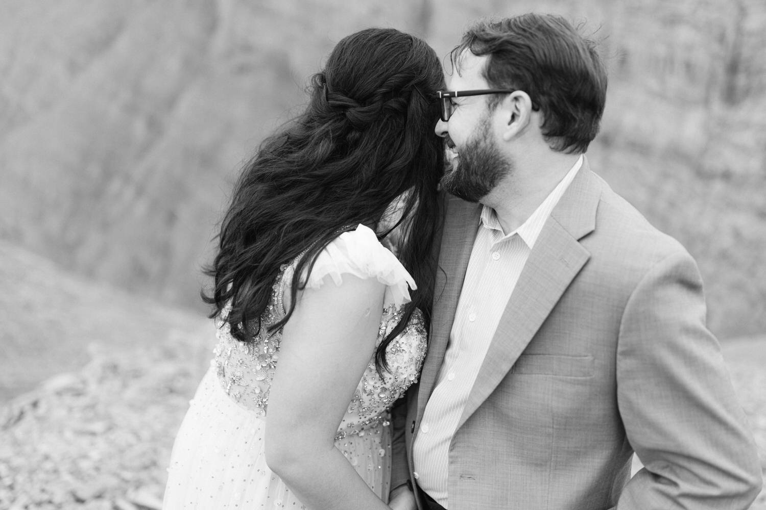 A couple sharing a loving moment, with the woman in a delicate dress and the man in a suit, captured in a black and white setting.