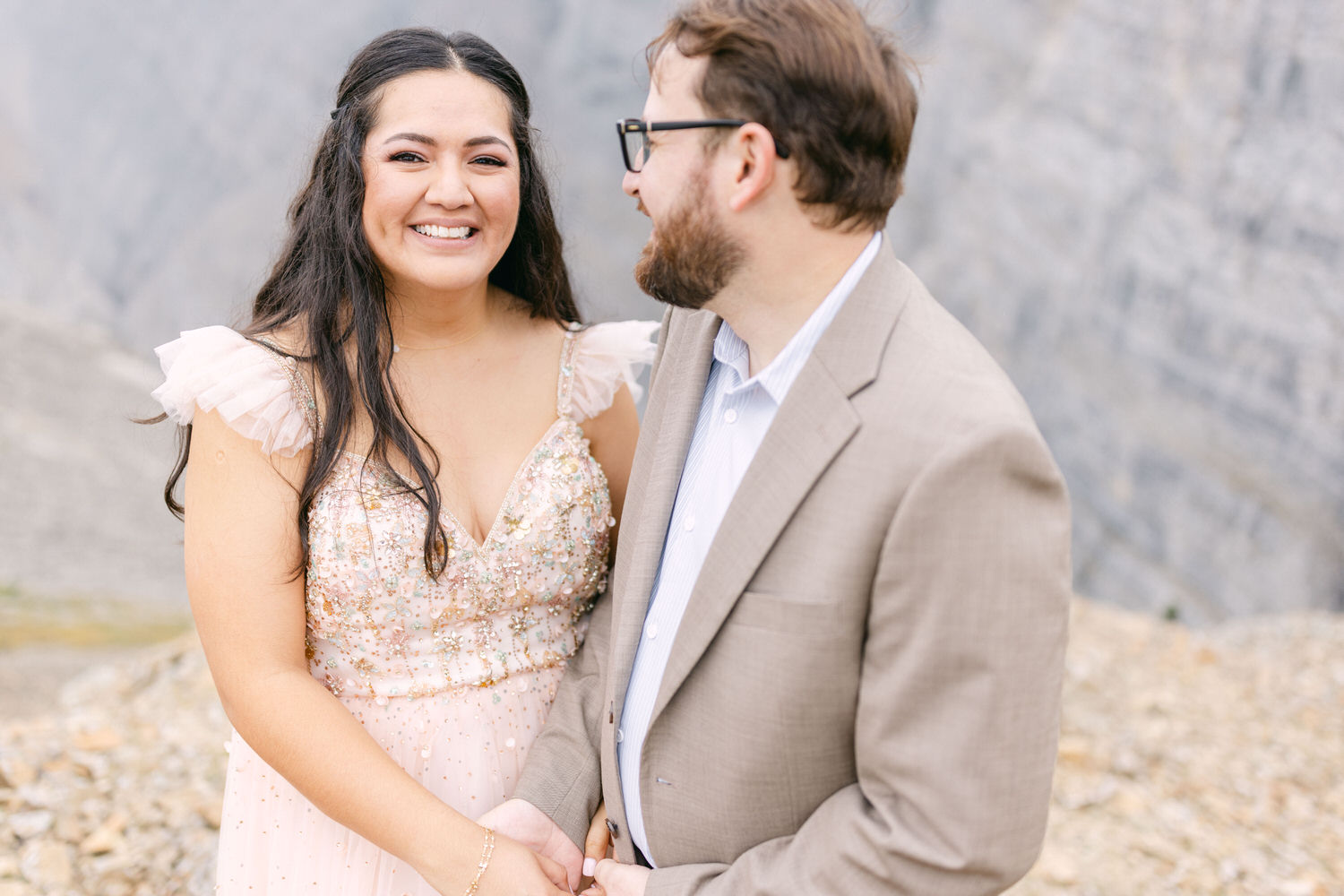 A smiling couple stands close together in a mountainous outdoor setting, with the woman wearing a sparkling, pale pink dress and the man in a light-colored suit, both looking at each other affectionately.