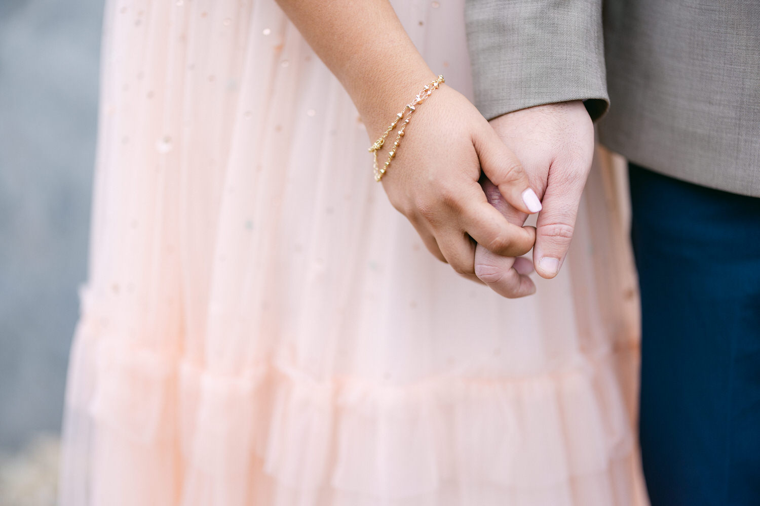 A close-up of two hands intertwined, showcasing a delicate bracelet on one hand and a soft, peach-colored dress in the background.