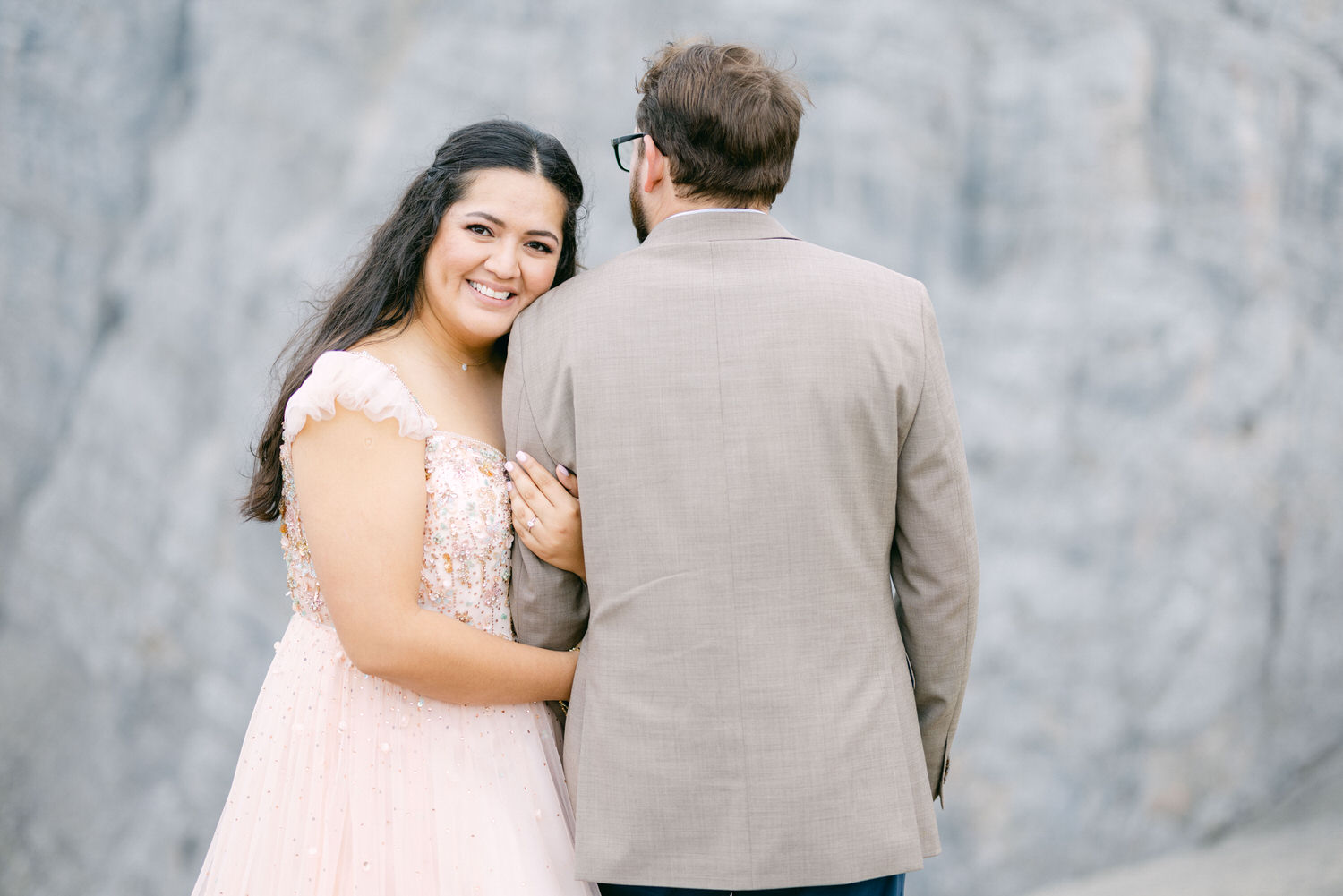 Joyful Couple Embracing::A smiling woman in a pink dress with sequins hugs a man in a light-colored suit, both enjoying a moment together against a soft, blurred background.