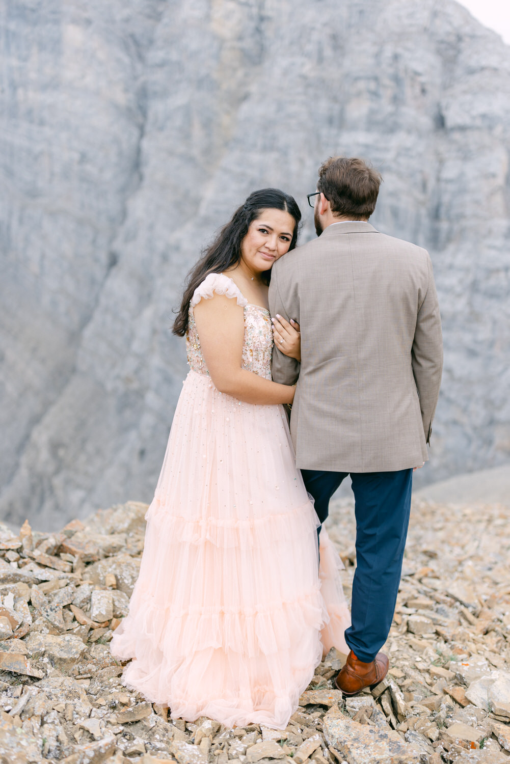 A woman in a pink dress smiles at the camera while embracing a man in a beige suit, set against a rocky mountain backdrop.