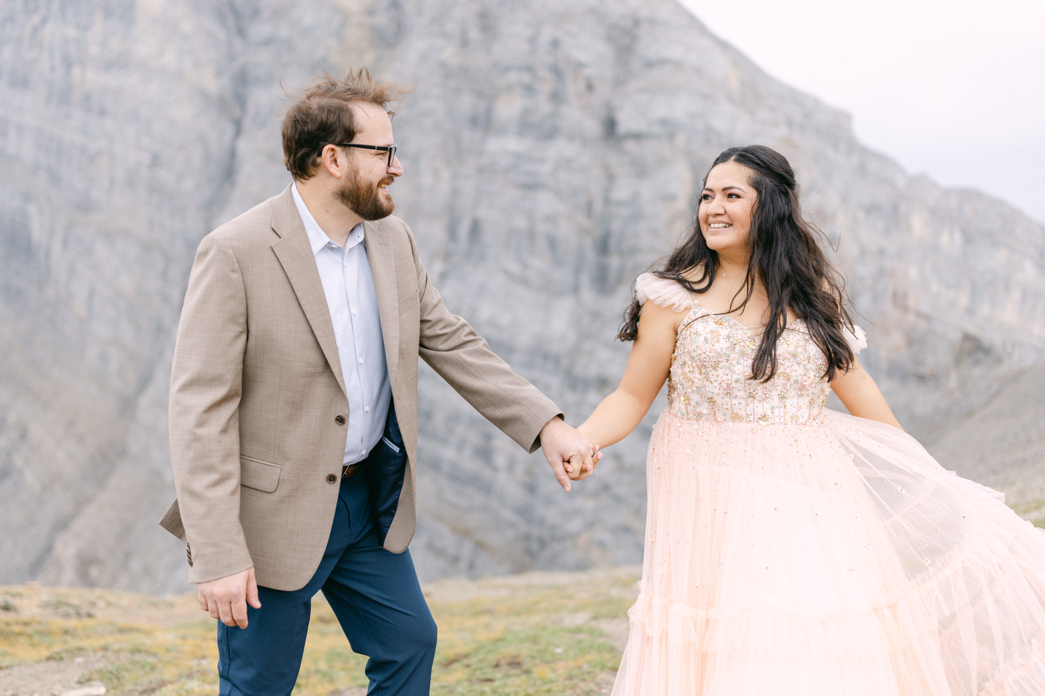 A couple sharing a joyful moment while holding hands in a scenic mountainous landscape, with the woman wearing a sparkling pink gown.