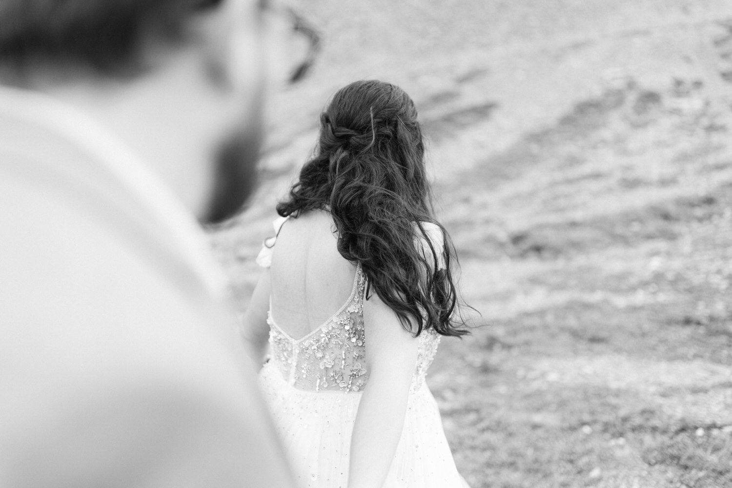 A bride in a sparkling dress turns away from the camera, with a gentle breeze catching her long, wavy hair, as a blurred figure gazes at her.