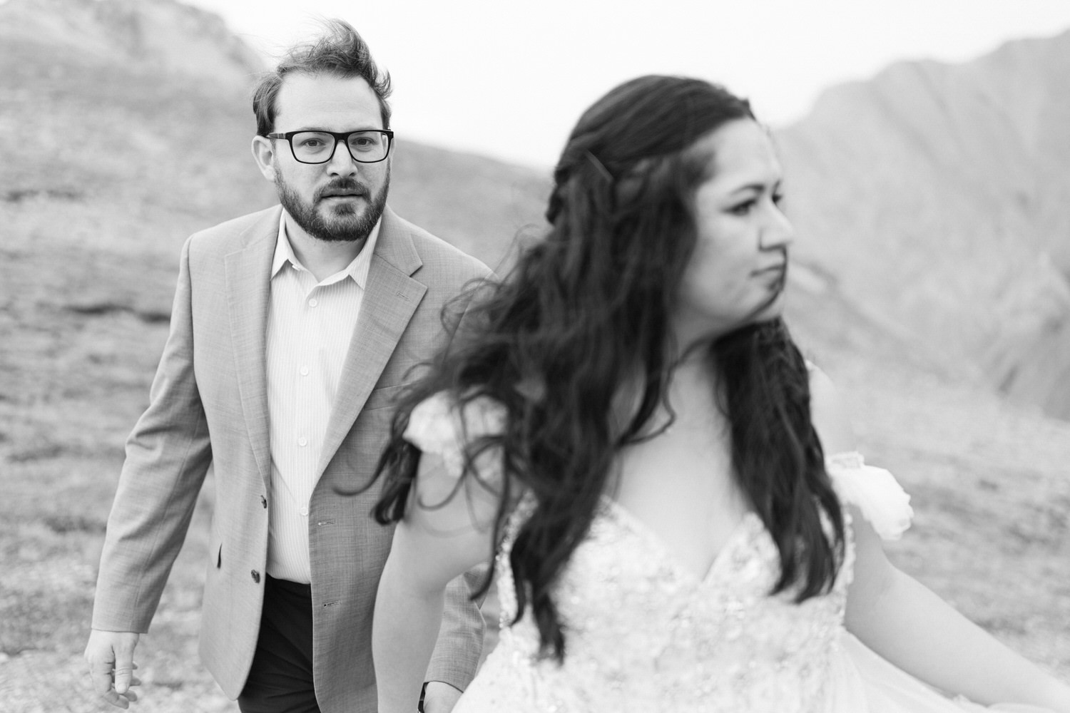 A bride in a flowing gown and a man in formal attire walk on a rocky terrain, capturing a tense or reflective atmosphere.