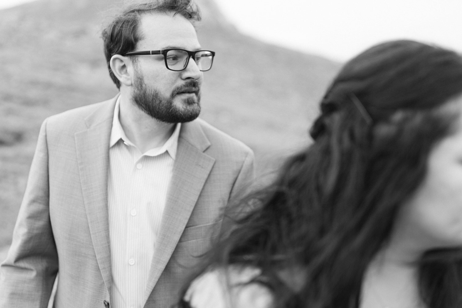 A candid black and white photograph of a man in a suit and glasses looking thoughtfully away from a woman whose face is partially visible, set against a blurred natural background.