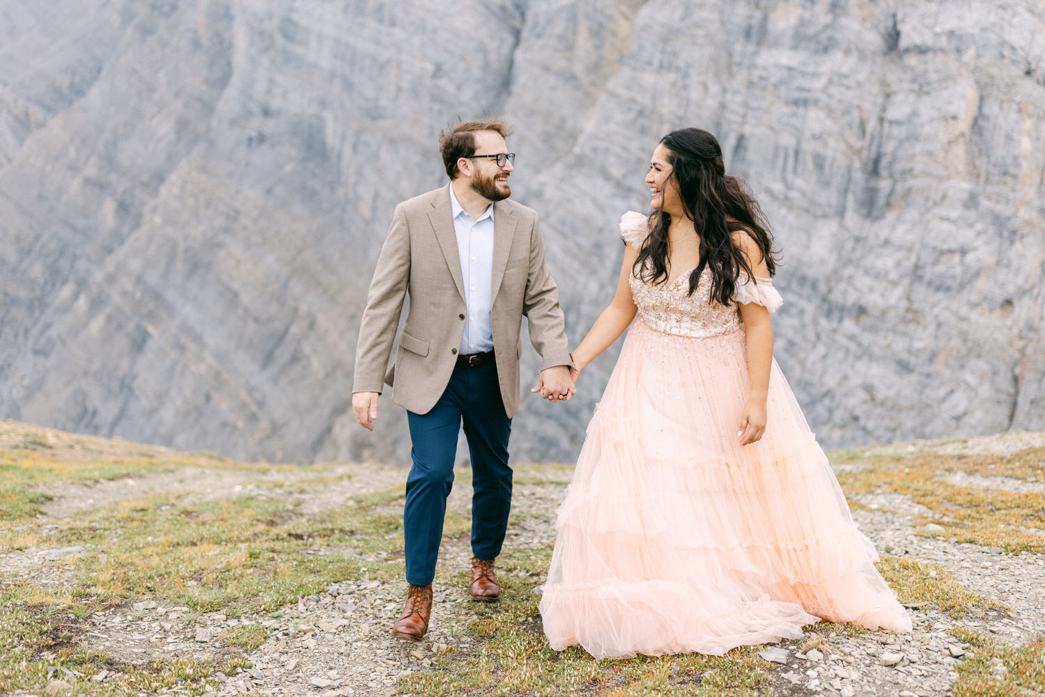 A couple enjoys a romantic moment while holding hands, set against a stunning mountainous backdrop. The woman wears a pink, flowing dress, while the man is dressed in a light blazer and smart casual attire.