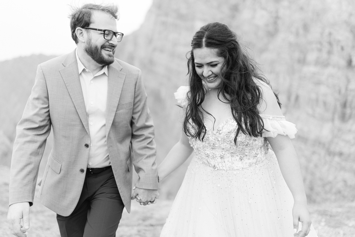 A happy couple holding hands and laughing while walking outdoors, dressed elegantly against a scenic backdrop.