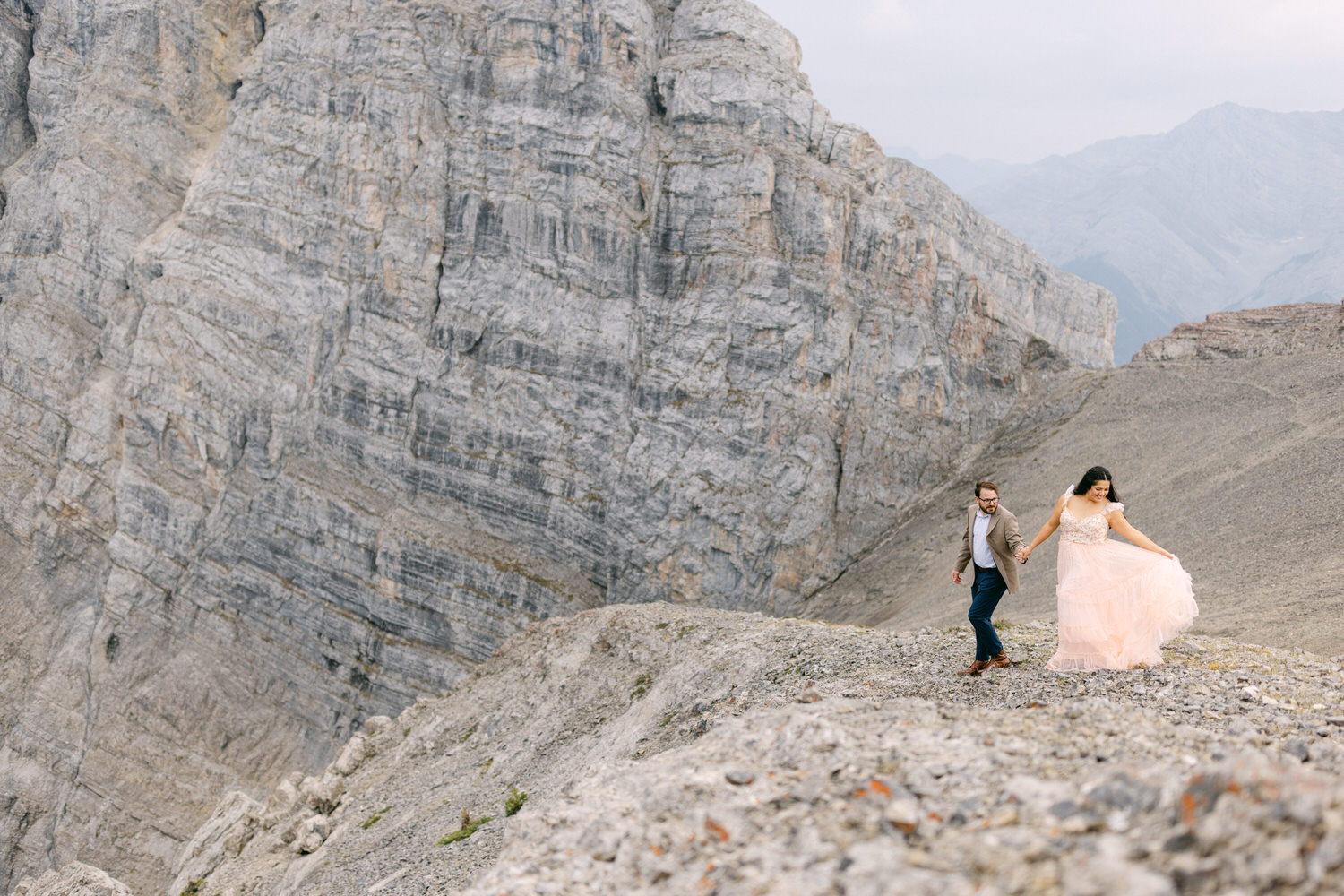 A couple joyfully walks hand in hand along a rocky mountain path, surrounded by a backdrop of towering cliffs and distant peaks, capturing a moment of love and exploration.