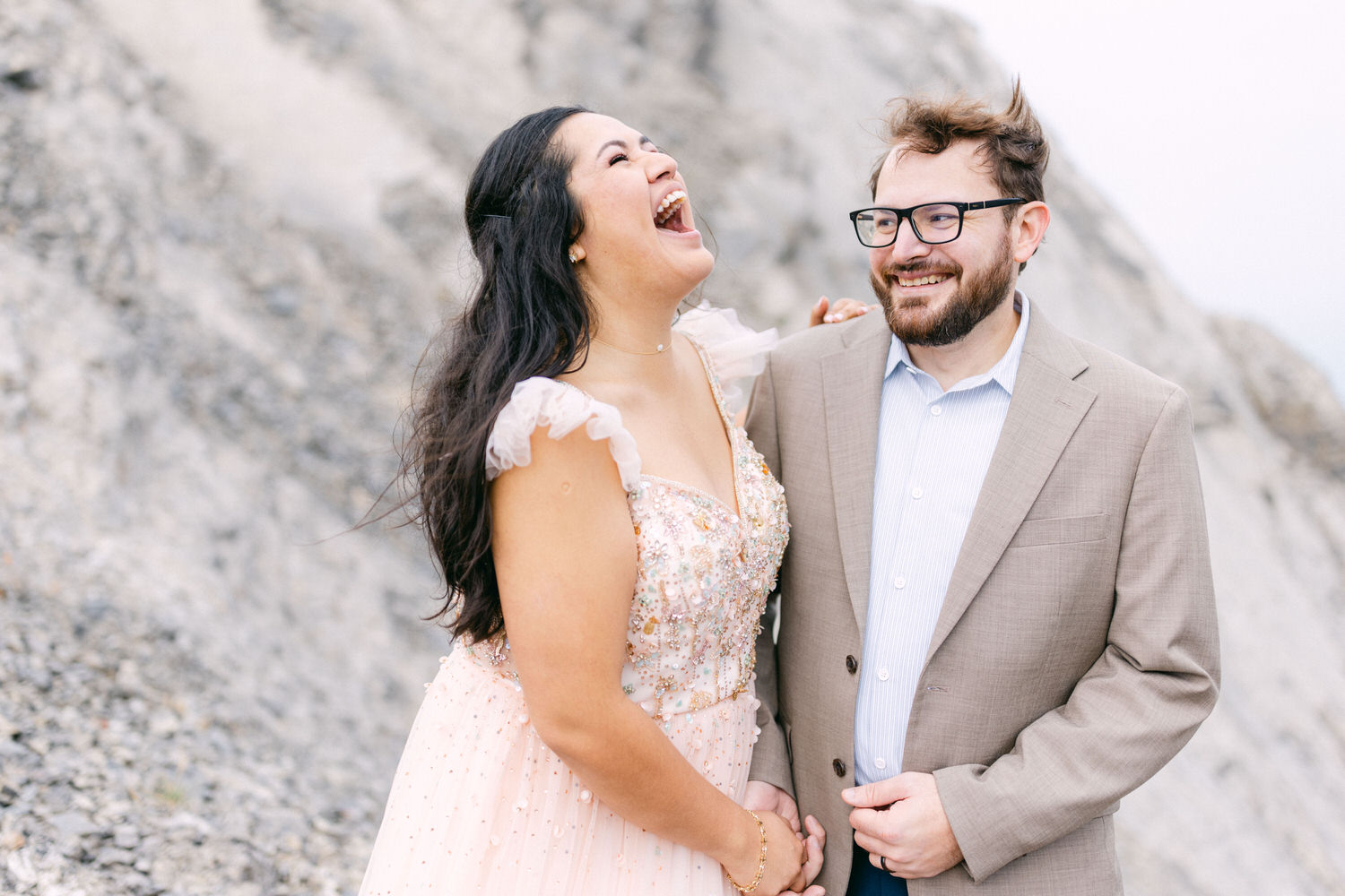 A couple laughs together outdoors, with the woman wearing a sparkling pink dress and the man in a tan suit, set against a natural rocky background.