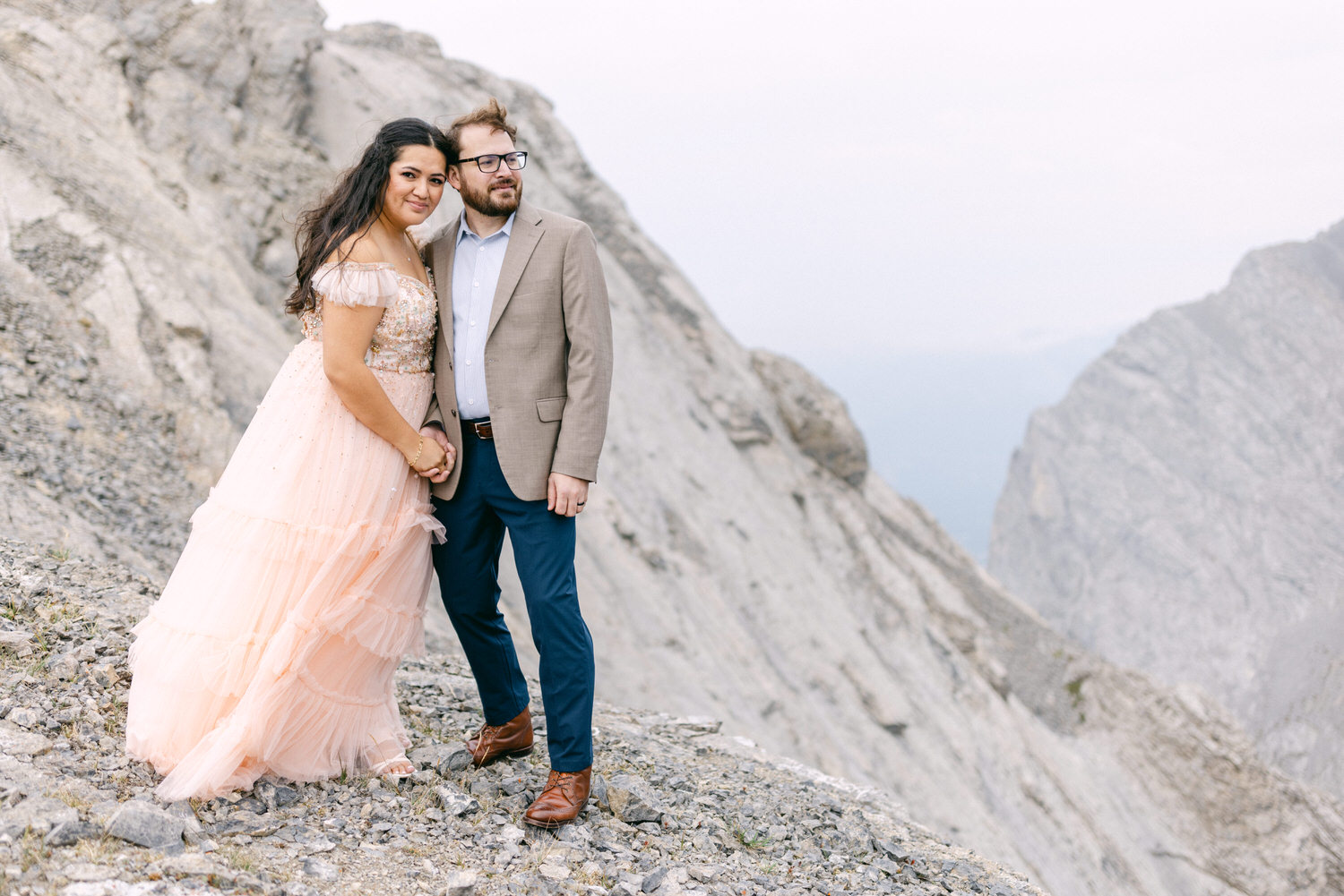 A couple stands together on a rocky mountain slope; the woman wears a blush pink gown while the man is in a light suit, both smiling gently.