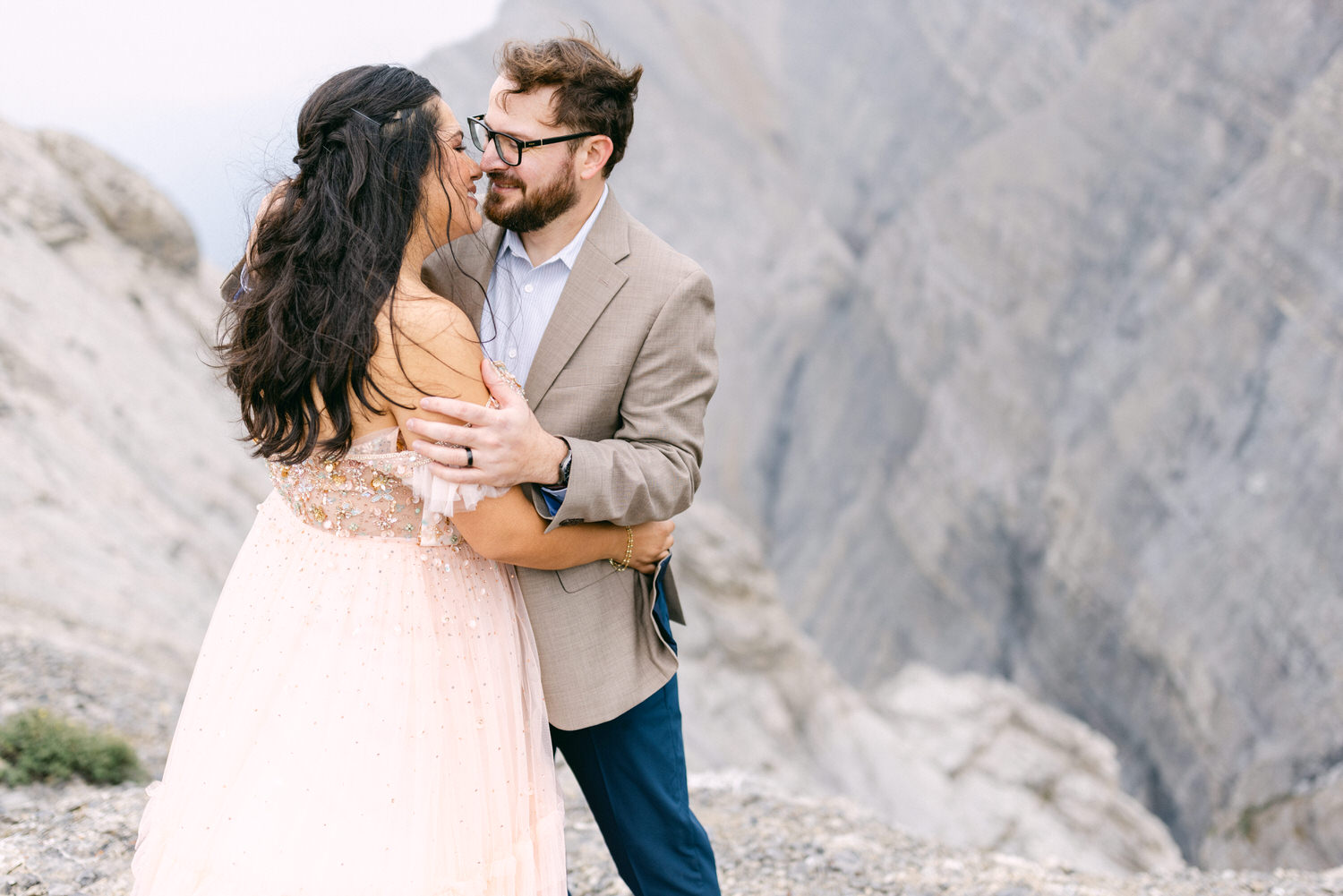 A man in a beige suit and a woman in a pink dress share a tender moment against a rocky backdrop, gazing into each other's eyes while holding each other closely.