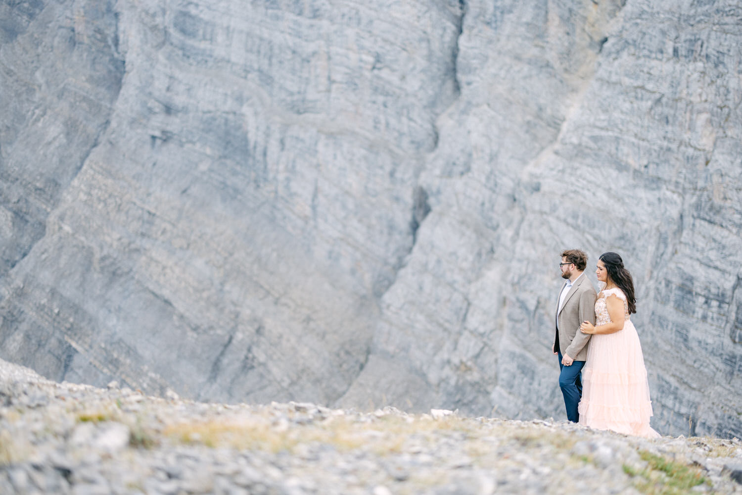 A couple in formal attire walks hand in hand along a rocky terrain, with a majestic, textured cliffside in the background.