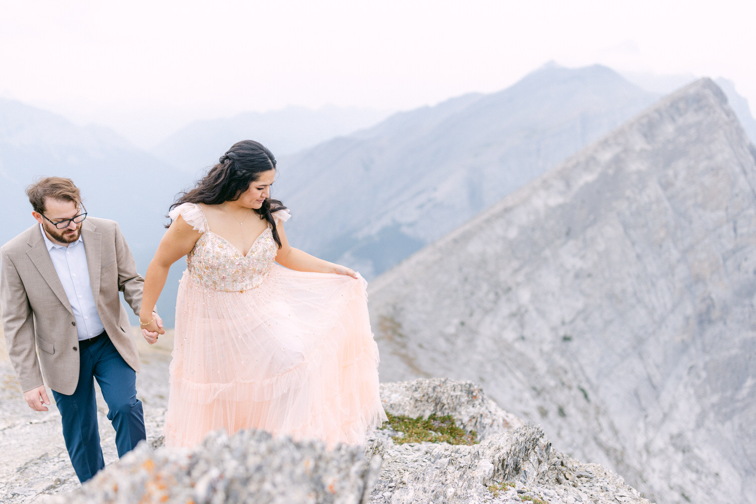 A couple walks hand in hand along a rocky mountain ledge, with the woman in a flowing pink dress embellished with sequins and the man in a formal blazer, surrounded by stunning mountain scenery.