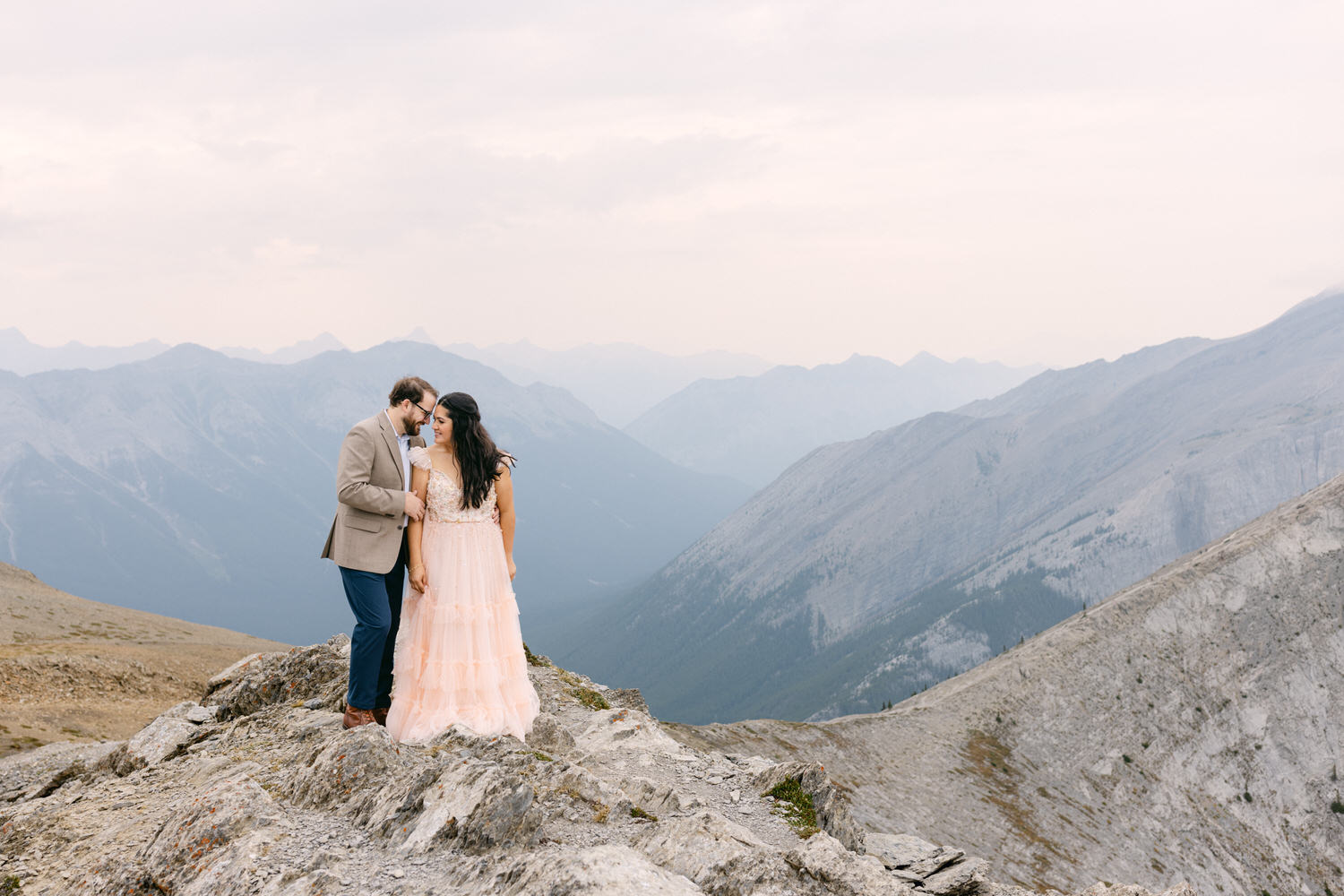 A couple sharing an intimate moment on a rocky mountain top, surrounded by a breathtaking landscape of distant mountains and a soft, cloudy sky.