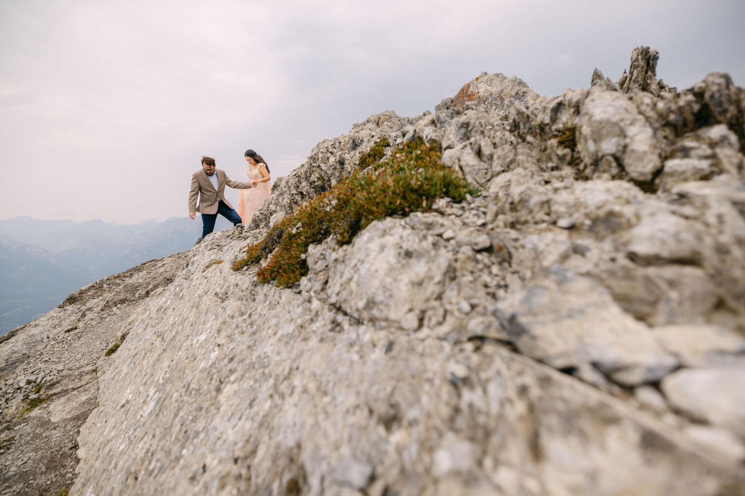 A couple in formal attire carefully ascending a rocky mountain, surrounded by a scenic landscape under a cloudy sky.