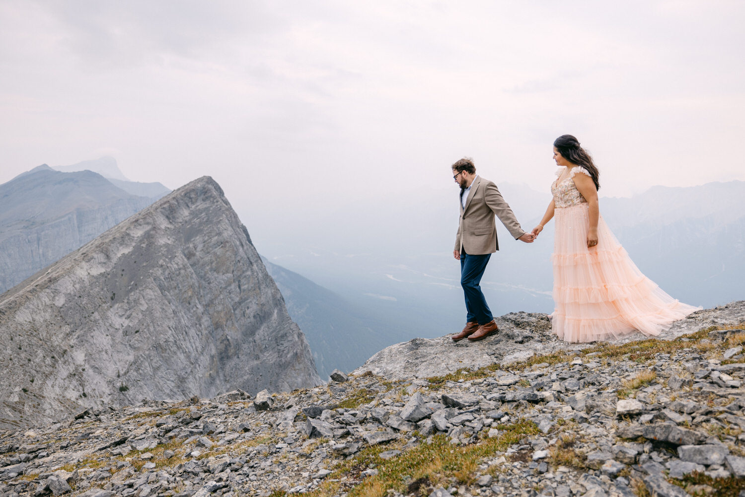 A couple in formal attire holds hands while standing on a rocky ledge with a dramatic mountain backdrop.