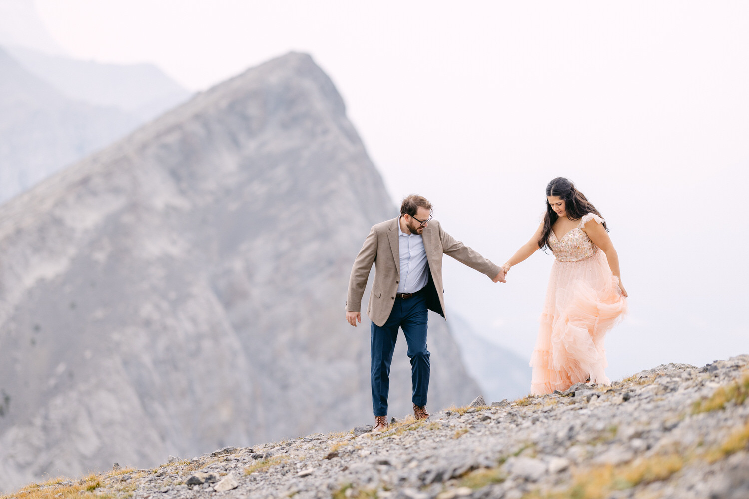 A couple holding hands as they walk along a rocky mountain trail, surrounded by rugged terrain and soft lighting.