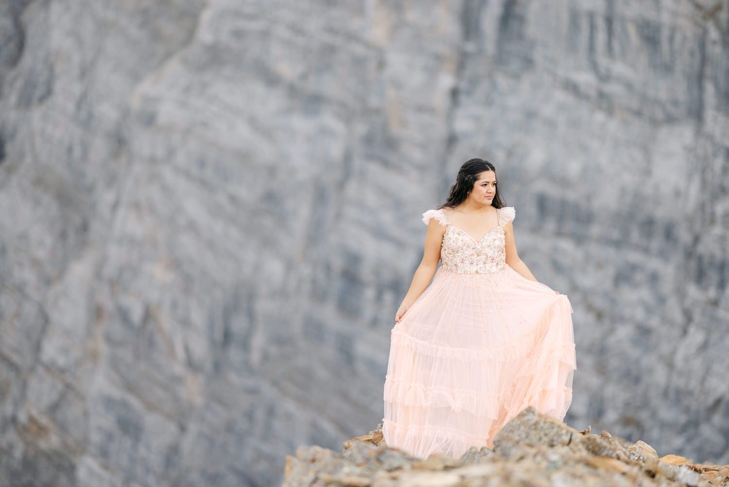 A woman in a flowing pink dress stands against a rugged rocky backdrop, showcasing a blend of delicate fashion and natural beauty.