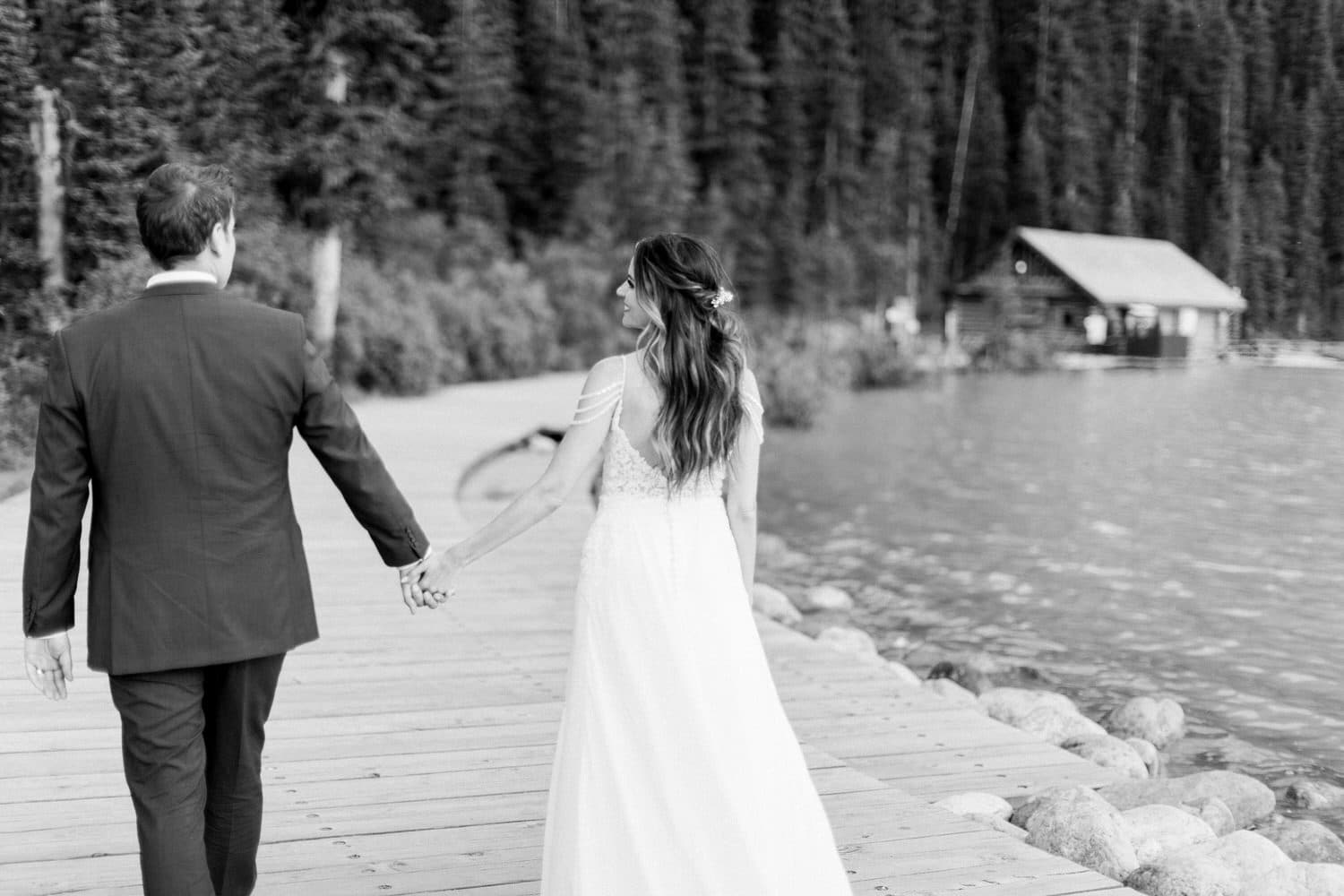 A couple walks hand in hand along a wooden path by a serene lake, surrounded by lush trees, capturing a moment of love and tranquility.