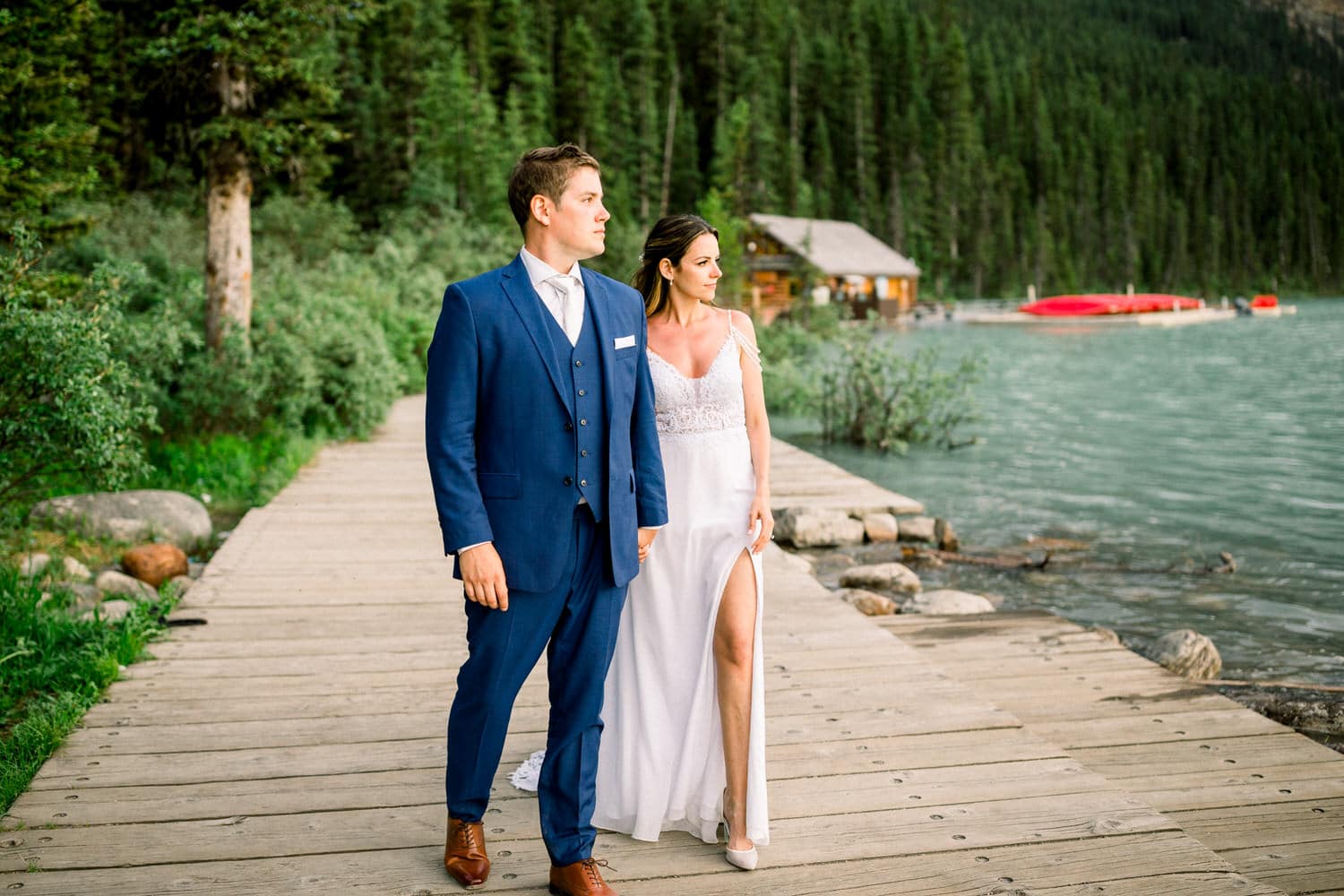 A bride and groom stroll hand in hand along a wooden path by a serene lake, surrounded by lush greenery and mountains.