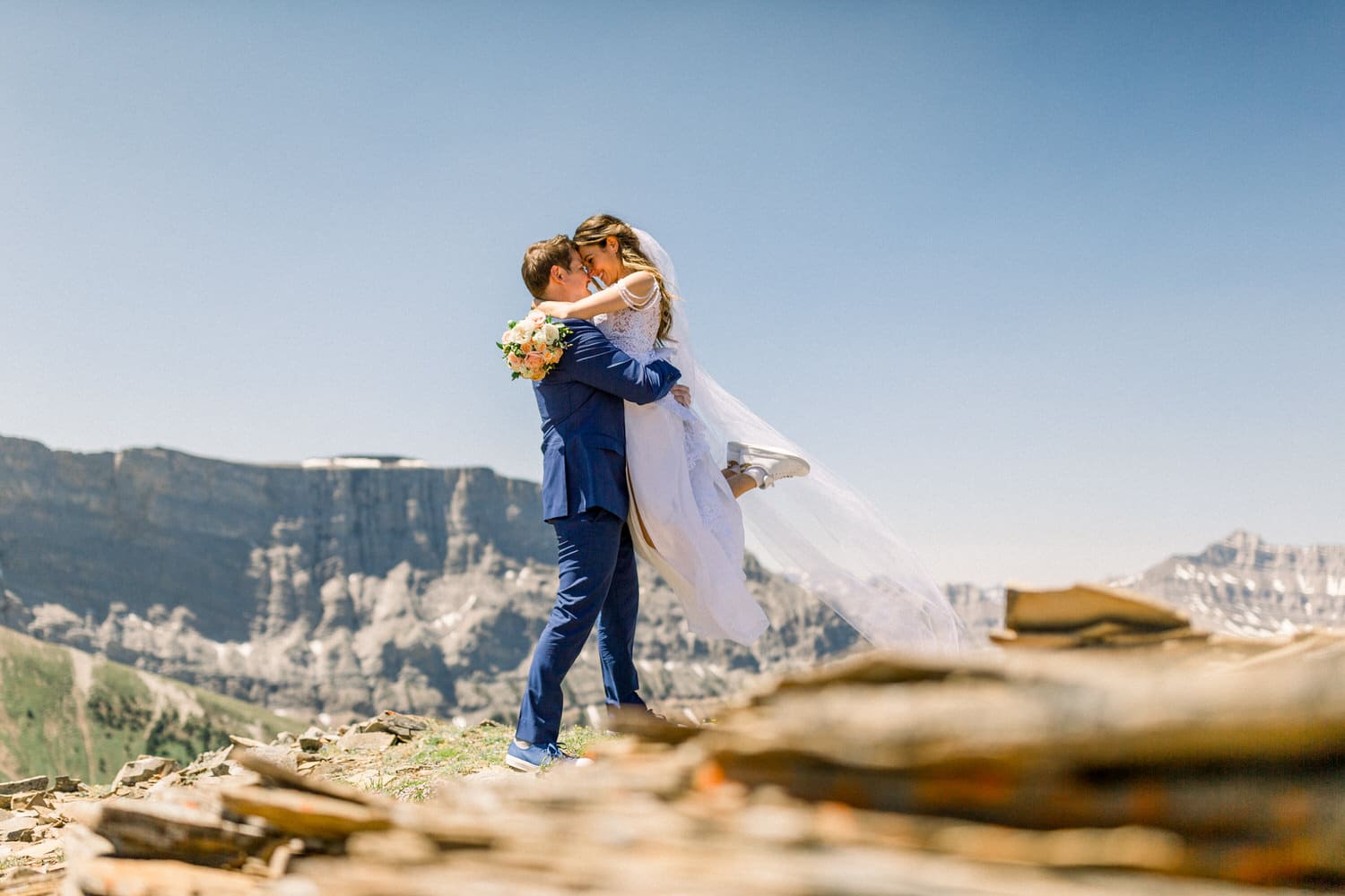 A newlywed couple shares a romantic embrace with a stunning mountain backdrop, as the bride holds a bouquet and wears a flowing veil.