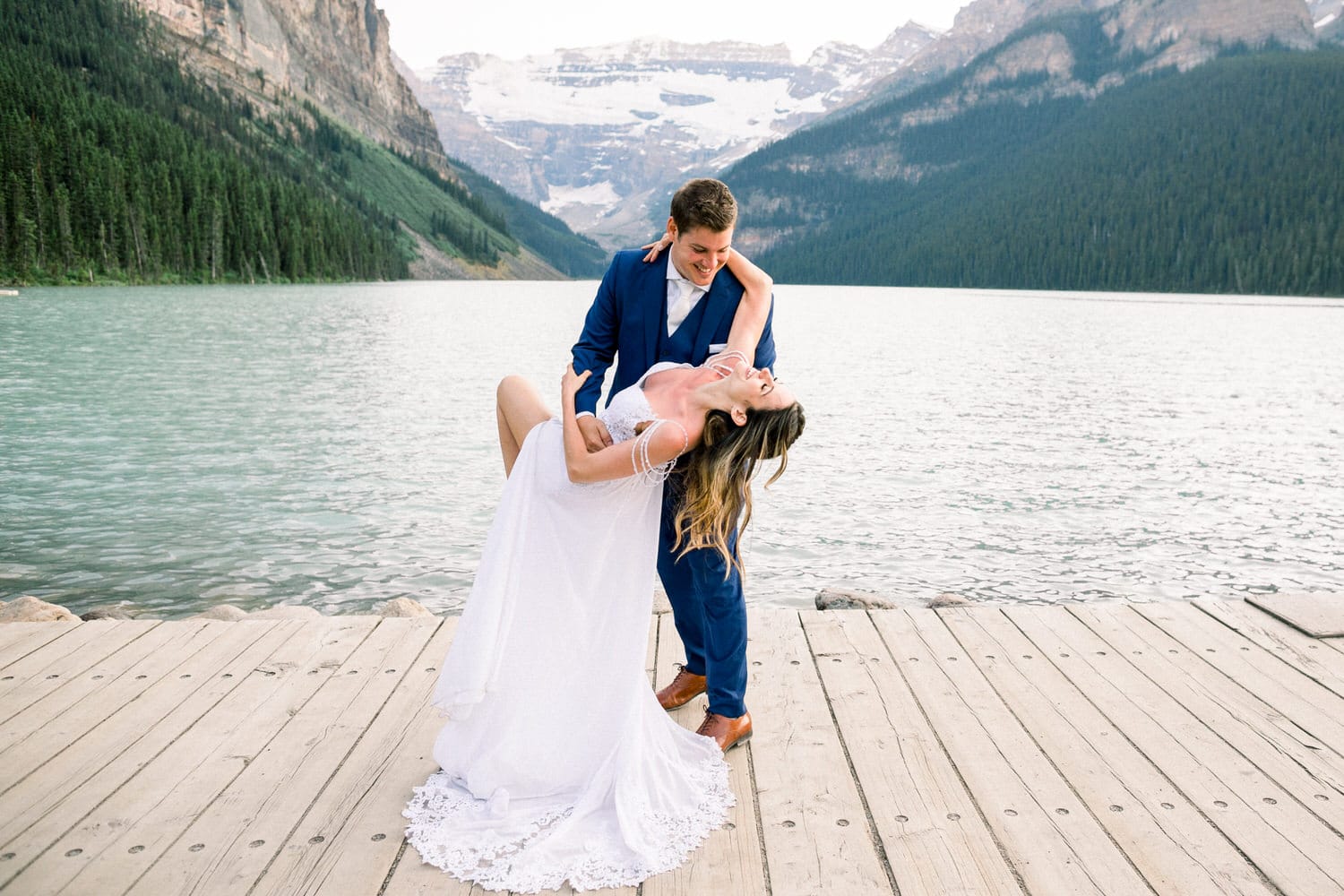 A couple in wedding attire joyfully posing on a dock by a lake surrounded by mountains, with the bride leaning back in a playful dip.