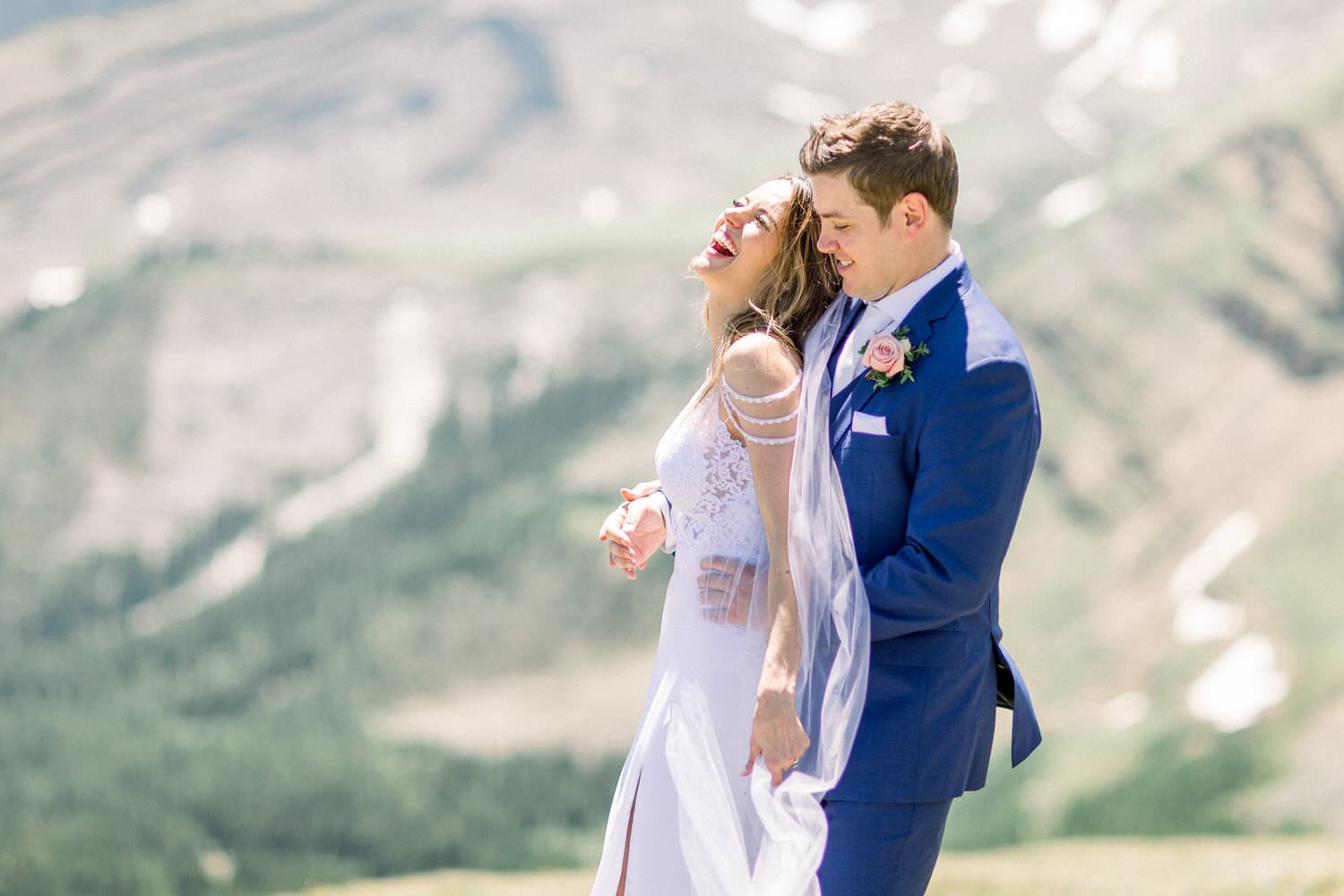 A couple shares a joyful moment in their wedding attire, surrounded by a breathtaking mountainous landscape.