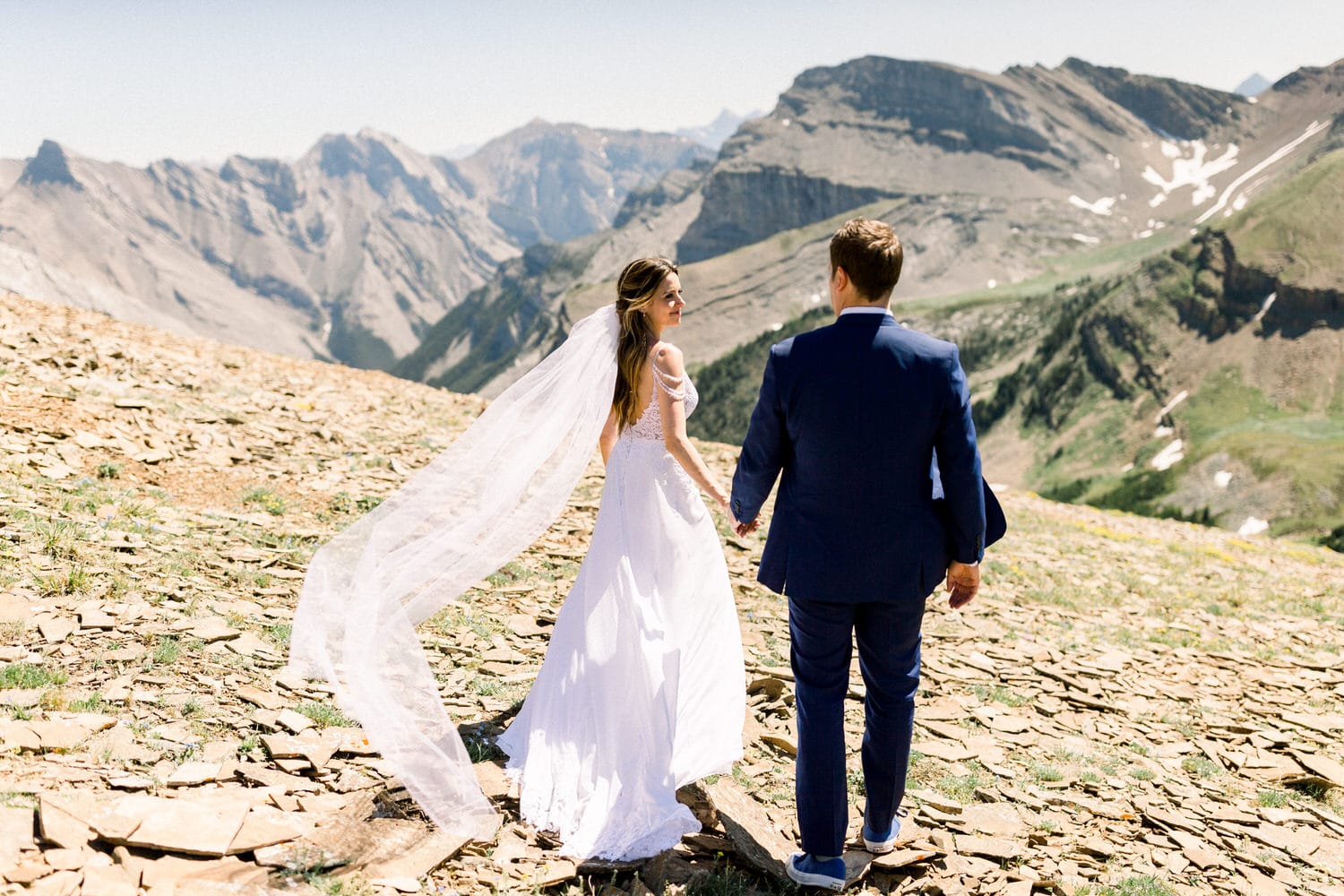 A couple walks hand in hand on a rocky mountain landscape, with majestic peaks in the background and a flowing veil in the breeze.