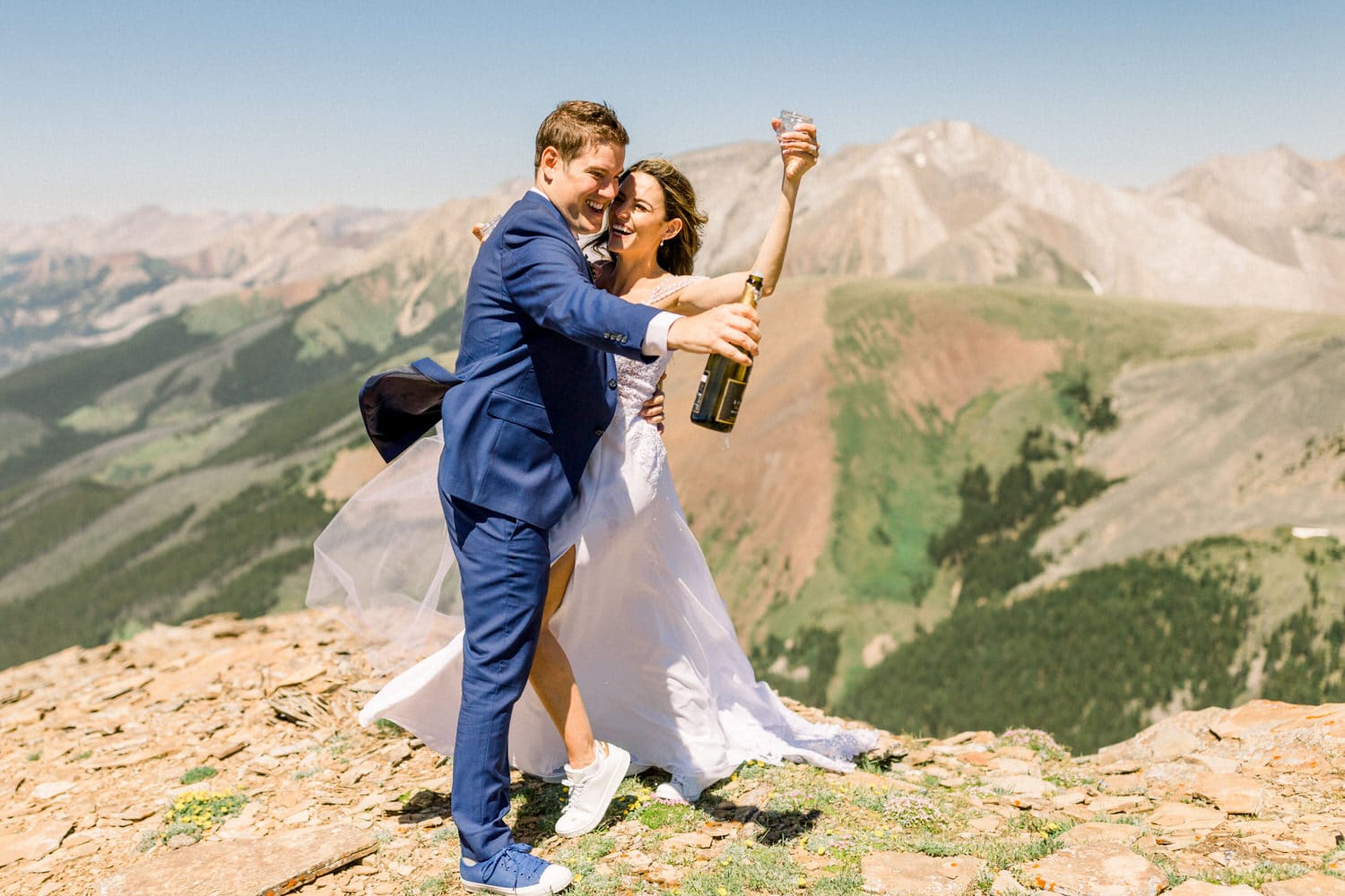 A joyful couple in wedding attire celebrating on a mountain peak, with drinks in hand and stunning mountain scenery behind them.