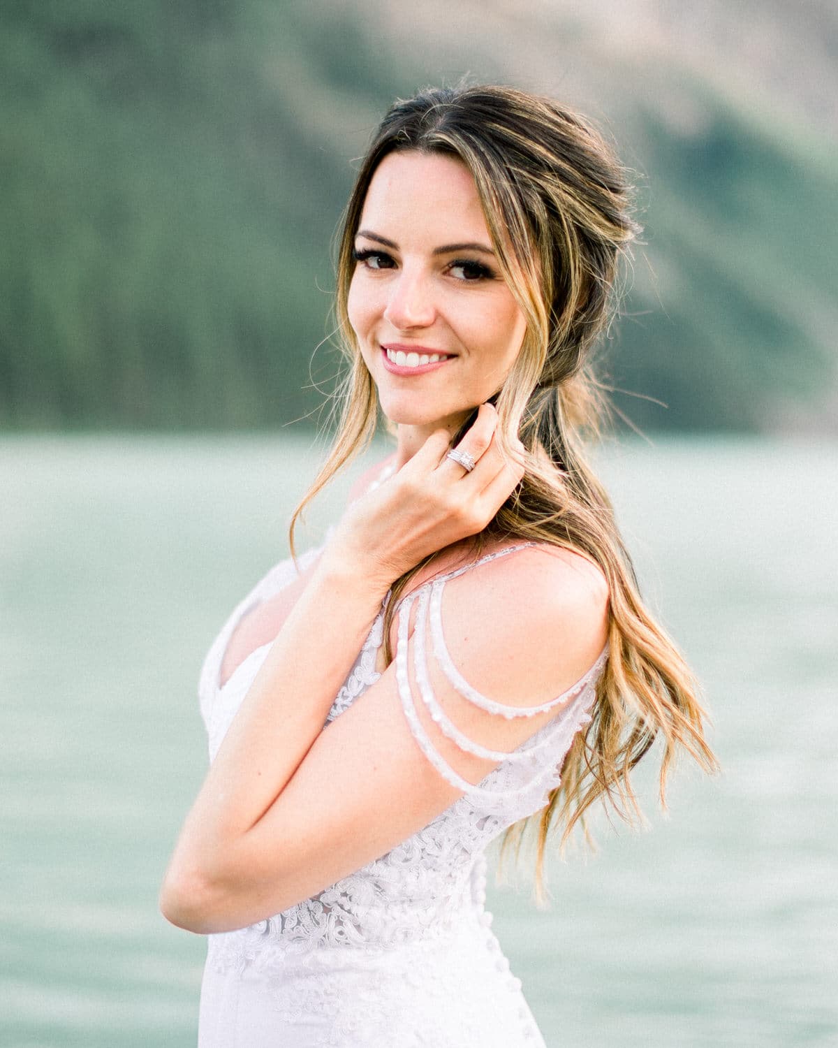 A smiling bride in a delicate wedding dress poses elegantly by a serene body of water, showcasing her engagement ring and sun-kissed hair.