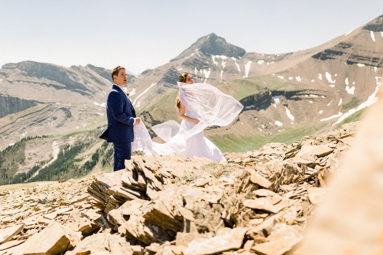 A couple in wedding attire stands on rocky terrain with a backdrop of stunning mountains, as the bride's veil billows in the wind.