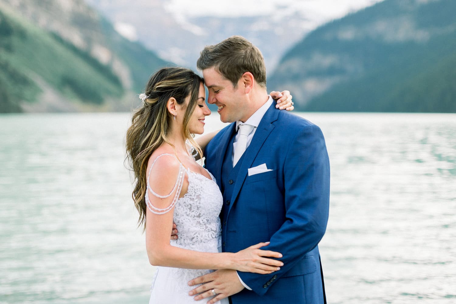 A couple sharing an intimate moment by a lake, surrounded by mountains, dressed elegantly for a special occasion.