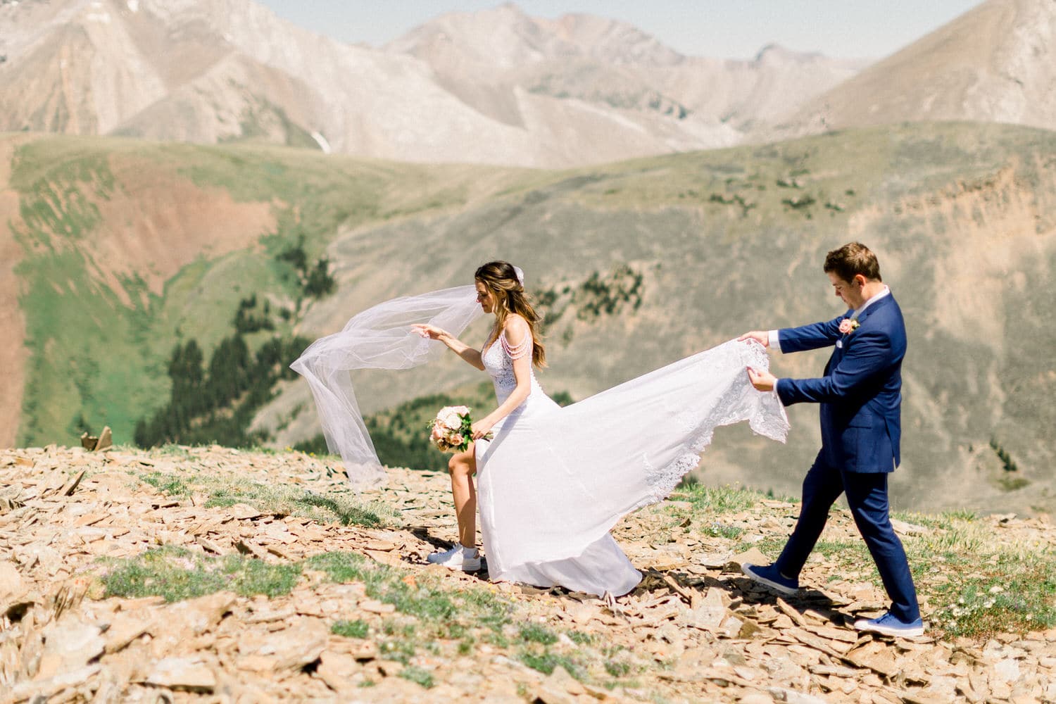 A bride in a flowing white gown and veil walks on rocky terrain as a groom assists her by holding her dress, set against a breathtaking mountain landscape.