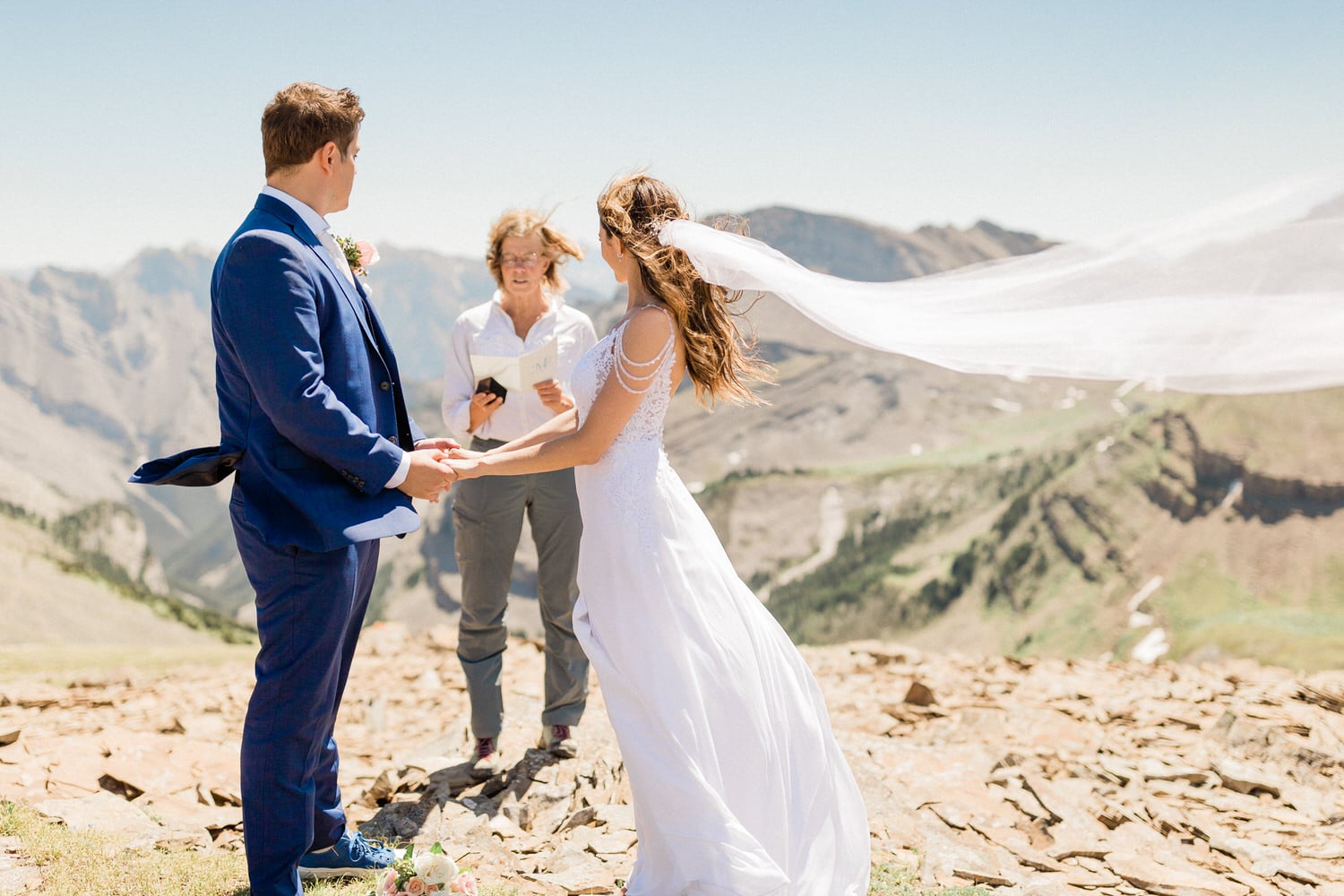 A couple exchanges vows during a scenic outdoor wedding ceremony, with a breathtaking mountain backdrop and a gentle breeze lifting the bride's veil.