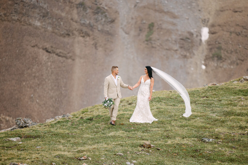 A bride and groom holding hands on a grassy hillside with a mountainous backdrop, capturing a moment of romance and joy.