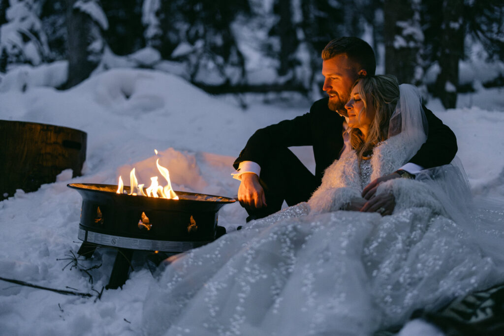 A couple in formal attire sits close together by a warm fire in a snowy landscape, capturing a moment of intimacy and romance.