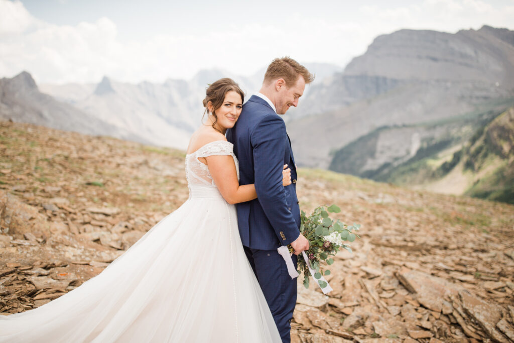 A groom and bride embrace in a picturesque mountain setting, with the bride in a flowing white gown and the groom in a navy suit, surrounded by stunning rocky landscapes.