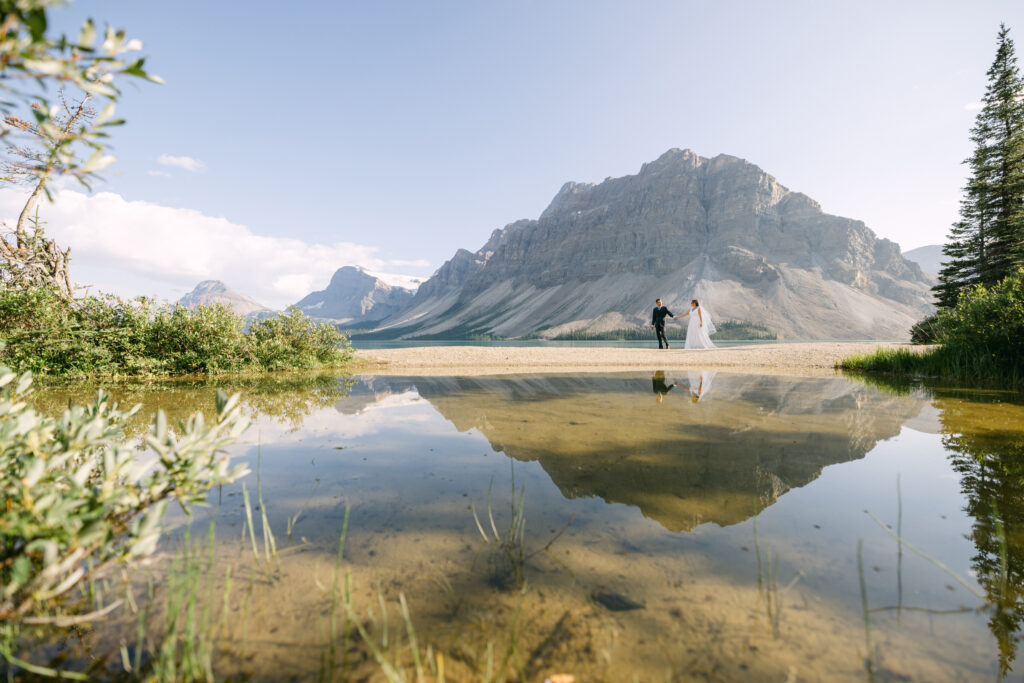 A couple in formal attire stands hand-in-hand by a serene lake, reflecting majestic mountains under a clear blue sky.