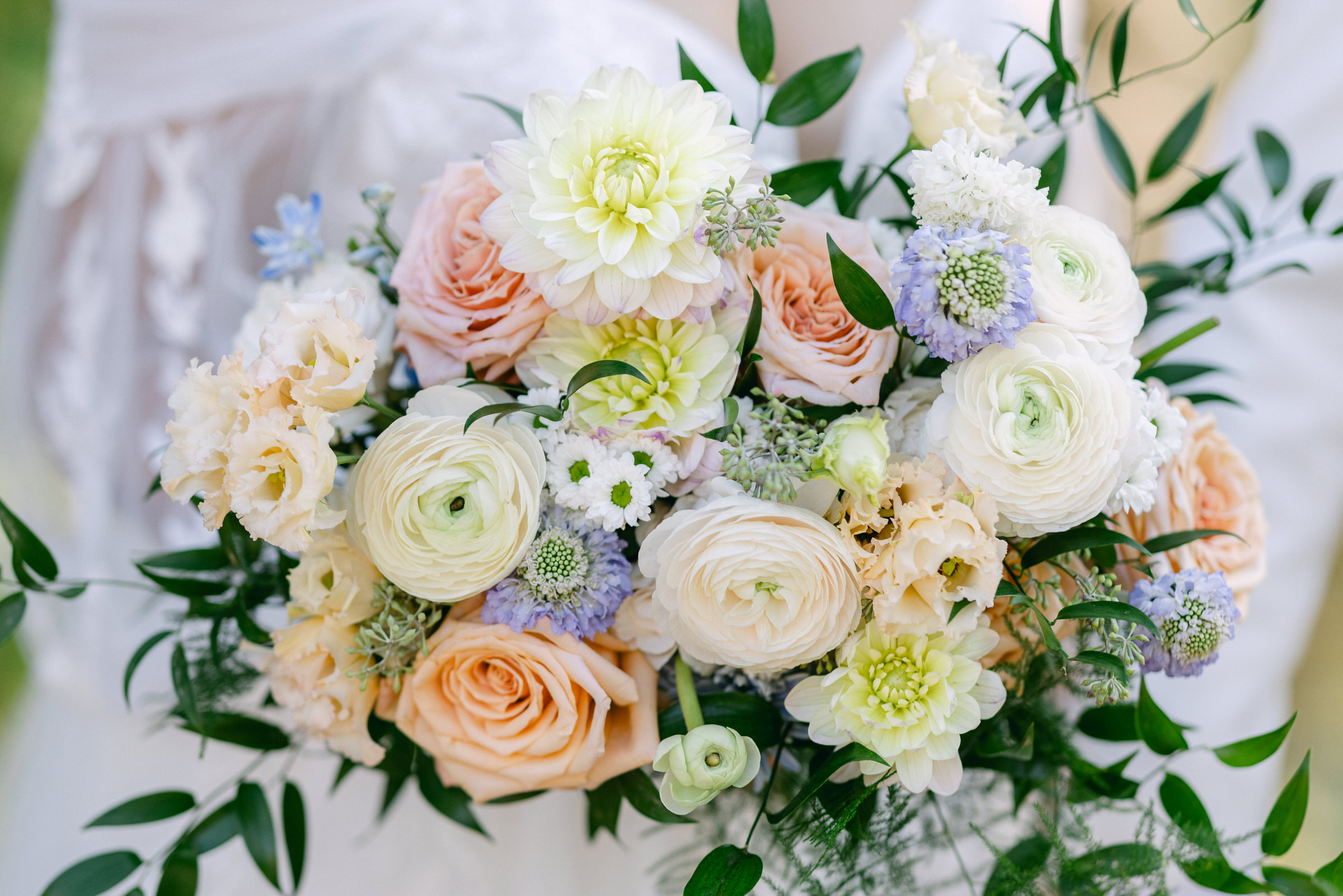 A close-up of a beautiful bouquet featuring pastel roses, ranunculus, and delicate greenery, held by a person in a soft-focus background.