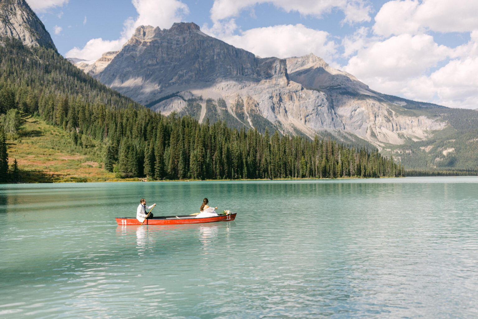 A couple paddling in a red canoe on a serene turquoise lake surrounded by lush forests and majestic mountains.