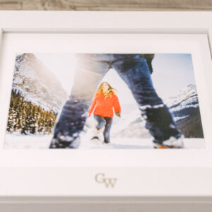 A framed photo of a woman in a red jacket walking through snow, with a distant mountain backdrop and a person's legs in the foreground.