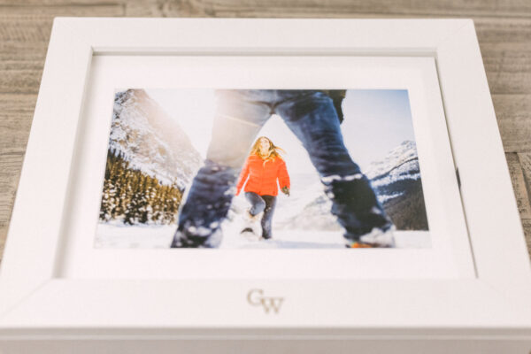 A framed photo of a woman in a red jacket walking through snow, with a distant mountain backdrop and a person's legs in the foreground.