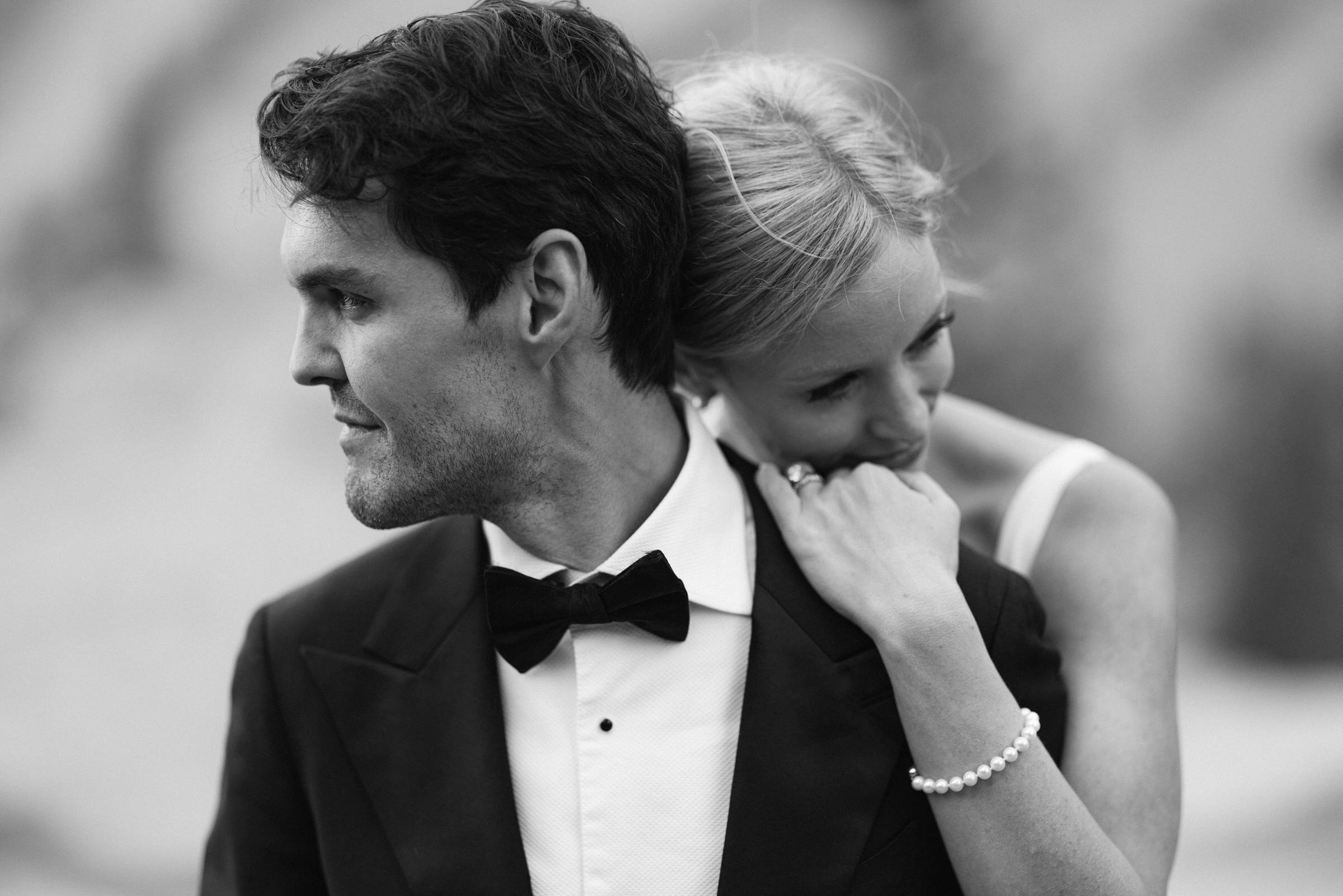 A groom in a tuxedo and a bride in a white dress share a tender embrace, with the bride resting her head on the groom's shoulder, captured in a soft black and white tone.